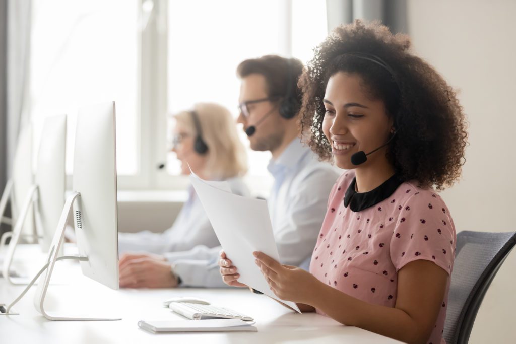 Smiling african american businesswoman call center operator agent wearing headset holding papers reading clients contacts working in customer service support helpdesk business office with colleagues